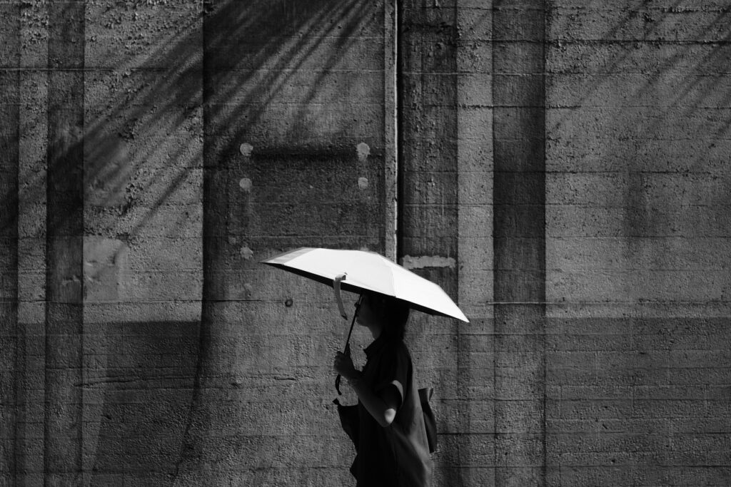 A person with a white umbrella walking beside a concrete wall, in black and white
