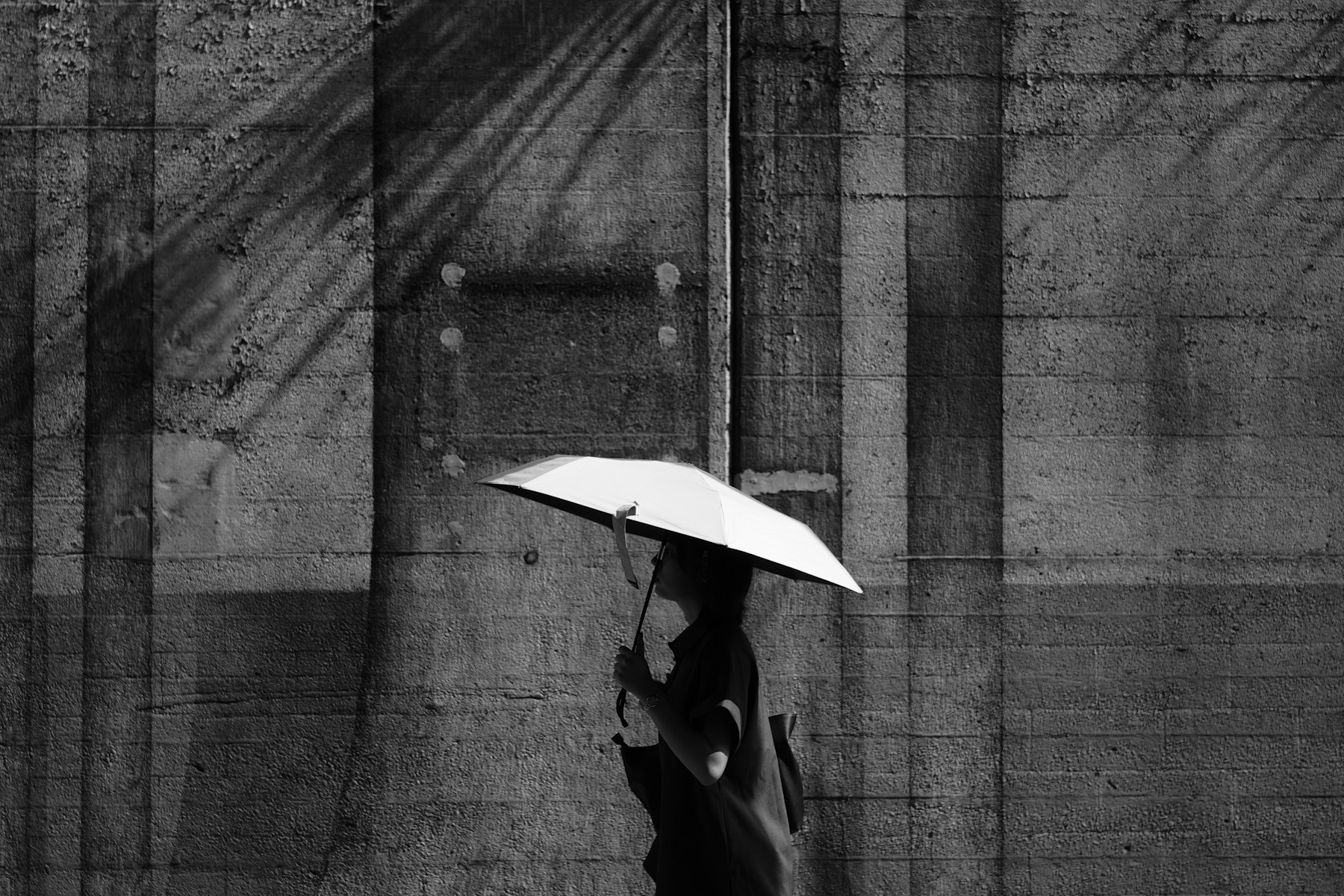 A person with a white umbrella walking beside a concrete wall, in black and white